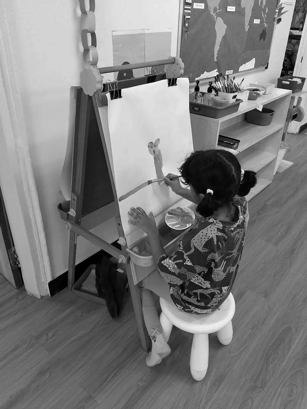 A young girl with glasses and pigtails paints a picture of a bear on a large white paper on an easel in a classroom. The classroom has shelves with art supplies and a world map on the wall.