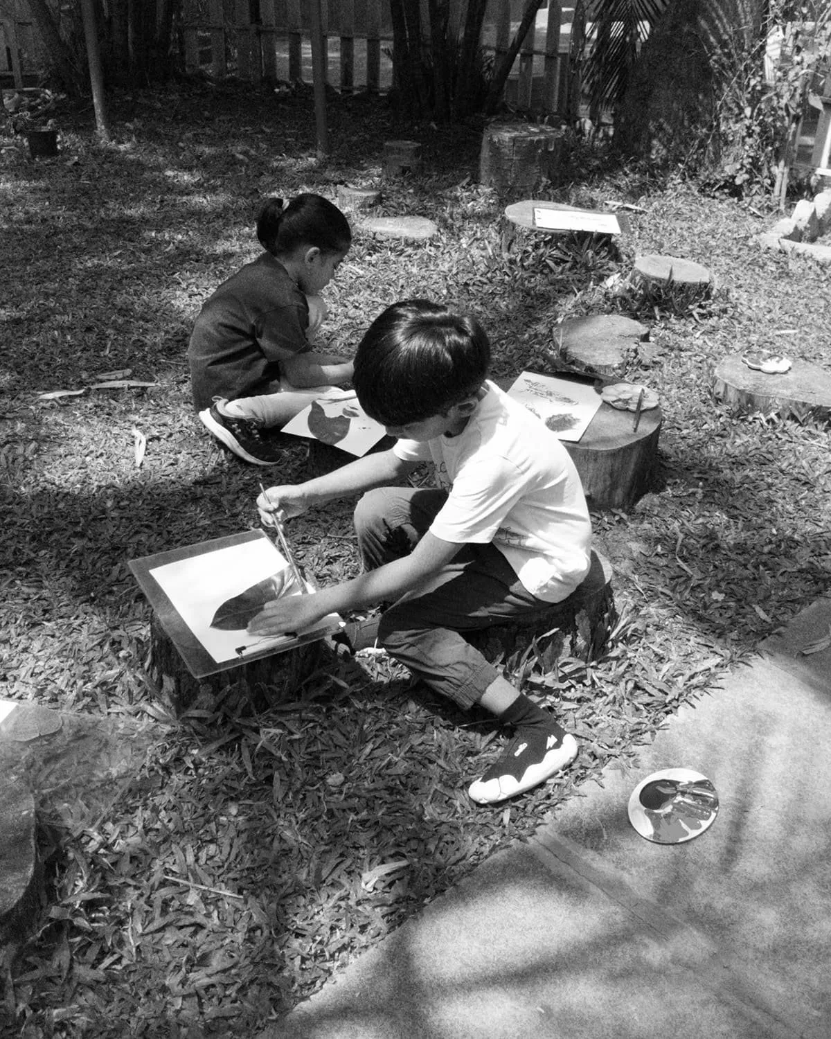 Two children sitting on tree stumps outdoors, painting pictures of leaves on paper, surrounded by leaves and natural elements.