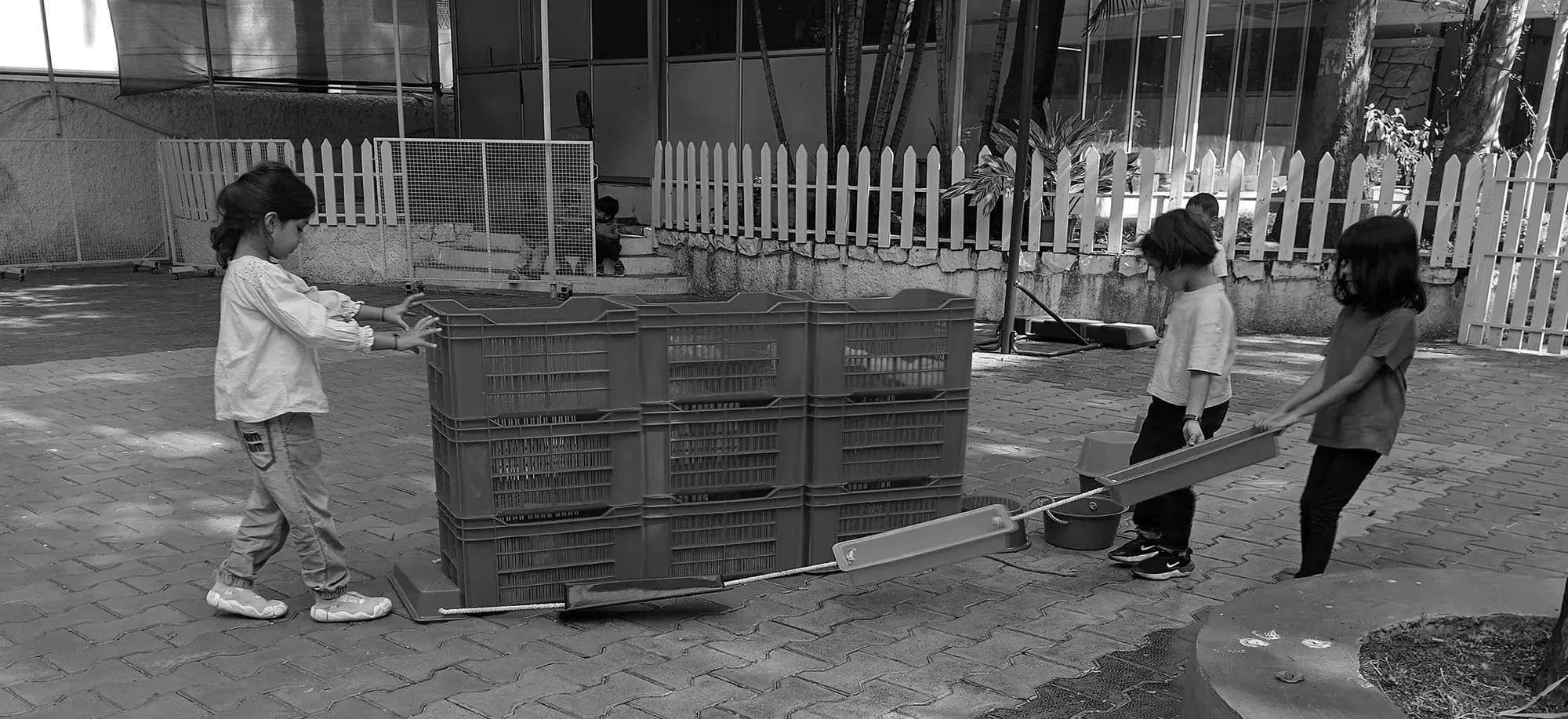 Four children playing outdoors with crates and a seesaw, some children sitting near the fence in the background.