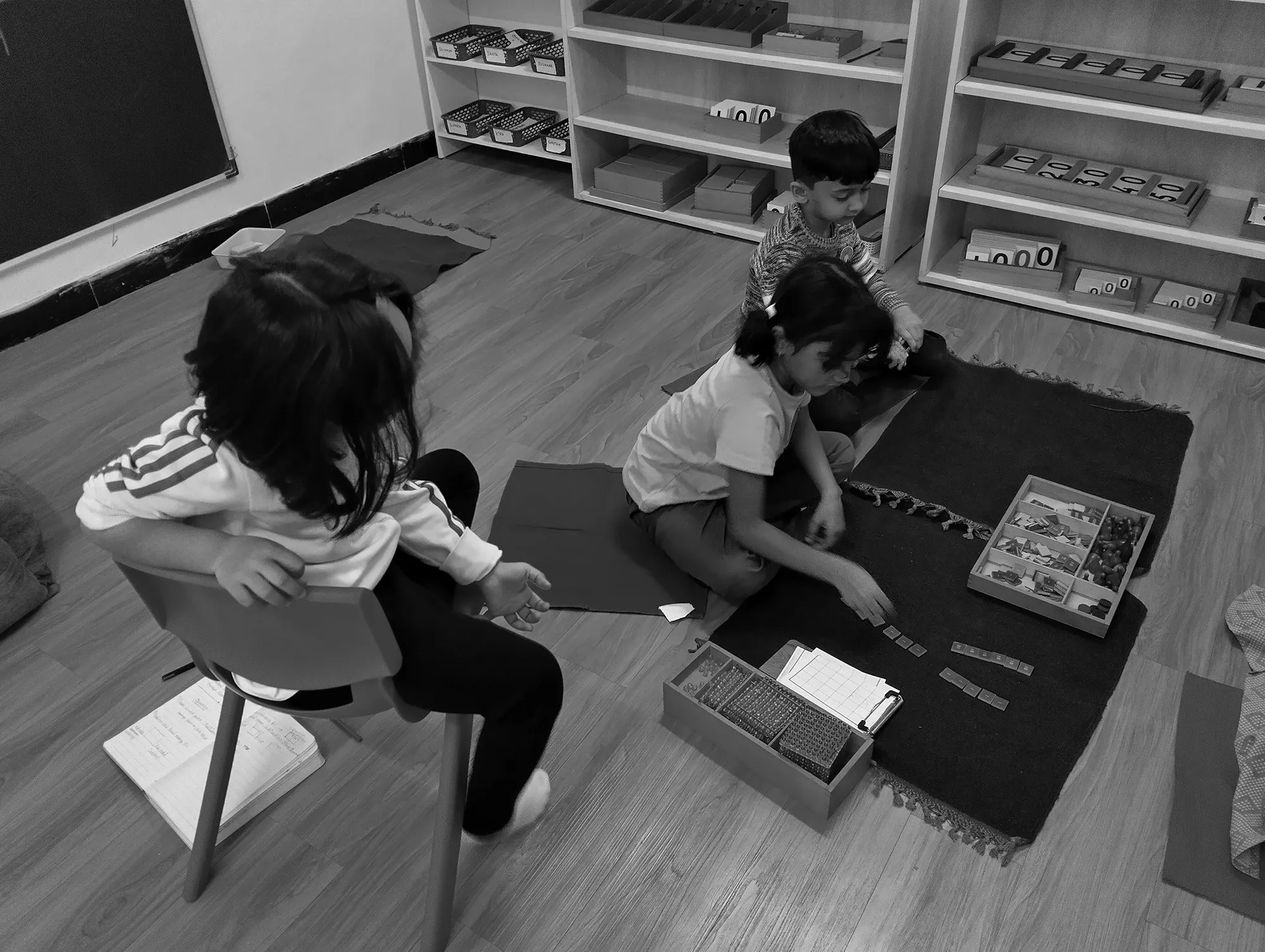 Three children sitting on the floor and a chair, engaging with educational toys and materials, in a classroom with shelves of educational items in the background.