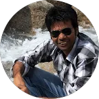 A man in sunglasses smiling while sitting near a waterfall or stream, with rocks in the background.