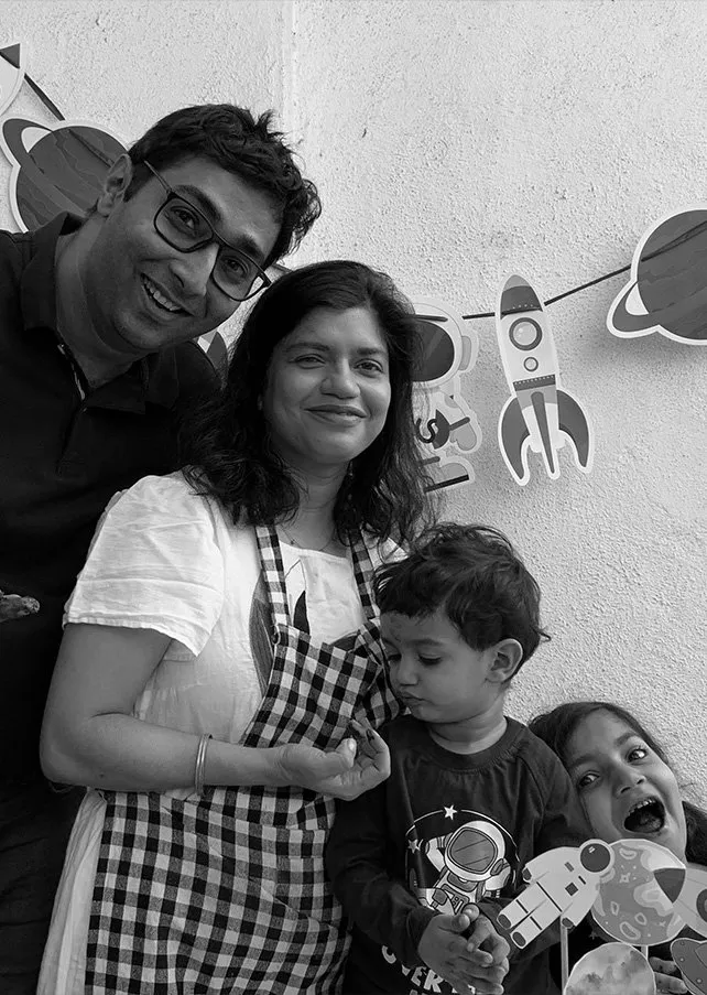 A family of four posing indoors with space-themed decorations in the background, including astronauts and planets. The adults are smiling, with one child looking down and another smiling widely.