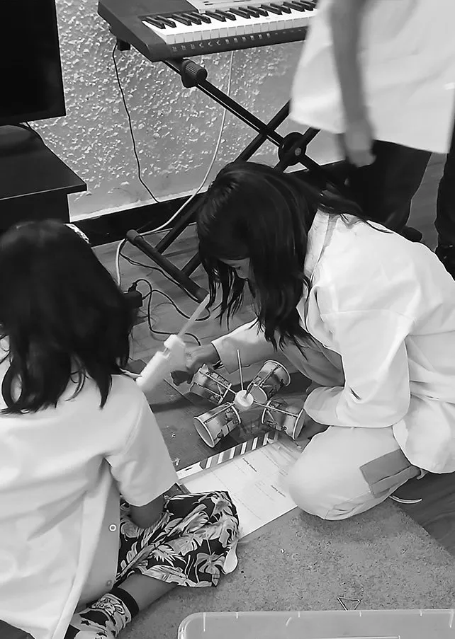 Two children in lab coats working on a small robotic spacecraft model on the floor, with a keyboard and microphone visible in the background.