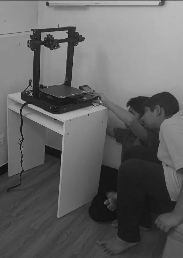 Two children are sitting on the floor next to a small printer and a 3D printer on a desk. One child is reaching for a remote control or computer mouse, and the other is leaning against the desk. The scene takes place in a classroom or office setting.