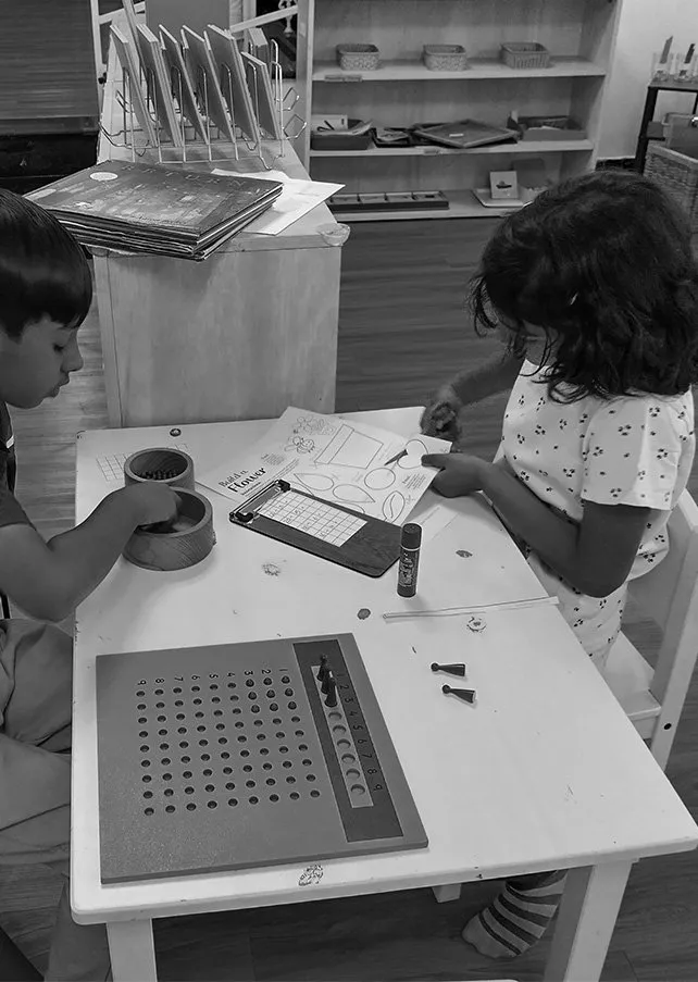 Two children sitting at a small table playing a board game, with various game pieces, a notepad, and markers. In the background, there is a wooden shelf with books and baskets.